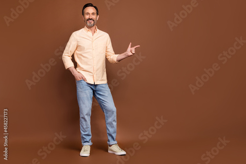 Photo of positive guy hand pocket direct finger empty space wear beige shirt jeans shoes isolated brown color background
