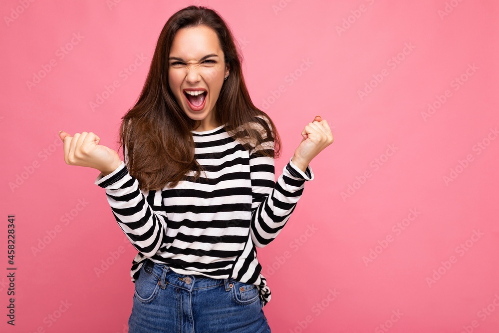 Portrait of young positive happy attractive brunette woman with sincere emotions wearing casual striped pullover isolated on pink background with empty space and celebrating victory shouting yeah