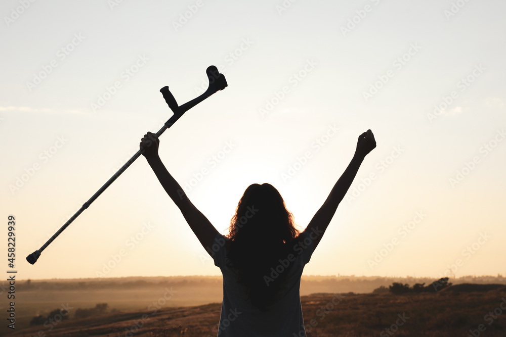 Fototapeta premium Woman holding elbow crutch outdoors at sunrise, back view. Healing miracle