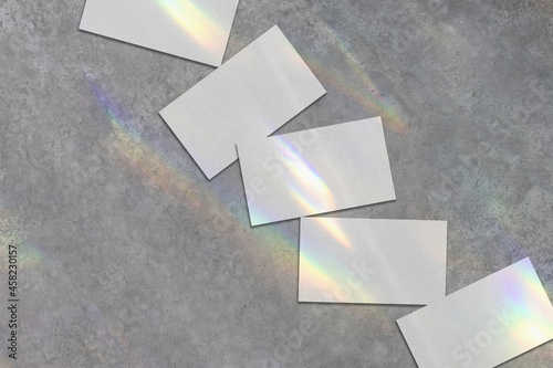 Close up of empty white rectangle business card mockups lying diagonally with rainbow crystal shadows and caustic light effect on light grey concrete background. Flat lay  top view. Open composition. 