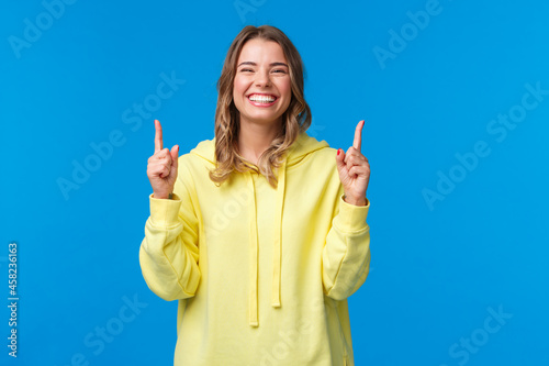 Cheerful happy smiling girl with blond hair in yellow hoodie, pointing fingers up and laughing carefree, recommend product, promo of subscribtion or company banner on blue background photo