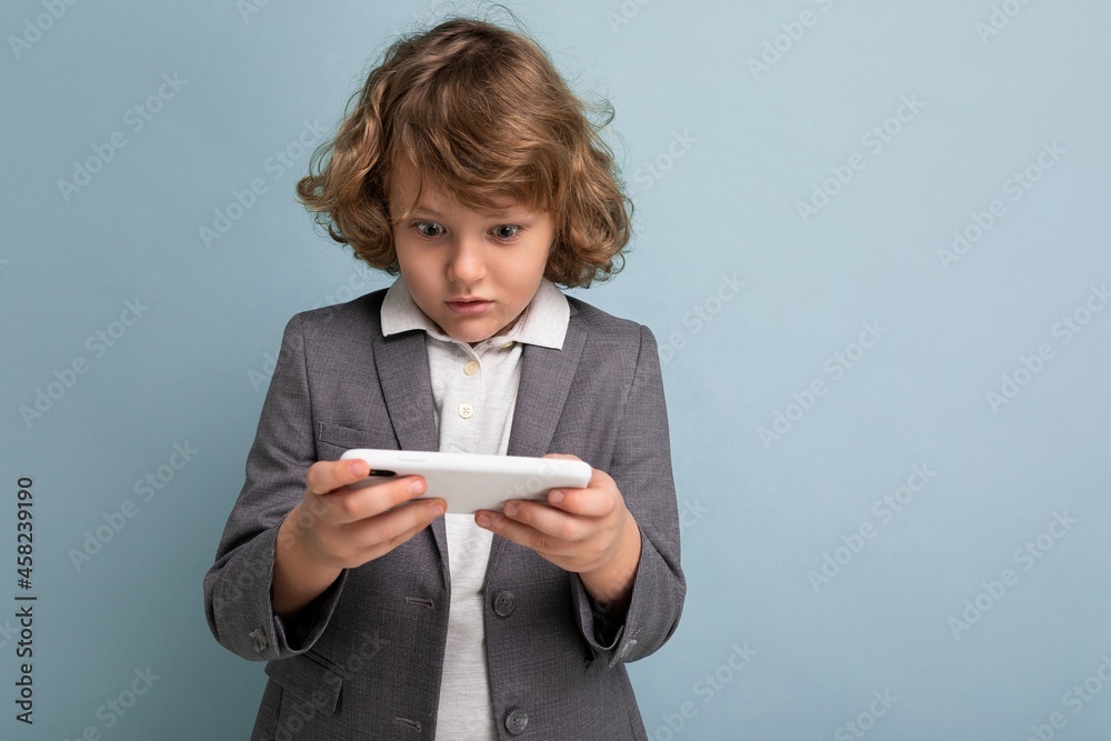 Photo shot of Handsome emotional child boy with curly hair wearing grey suit holding and using phone isolated over blue background looking at smartphone playing games Photo shot of Handsome emotional child boy with curly hair wearing grey suit holding and using phone isolated over blue background looking at smartphone playing games