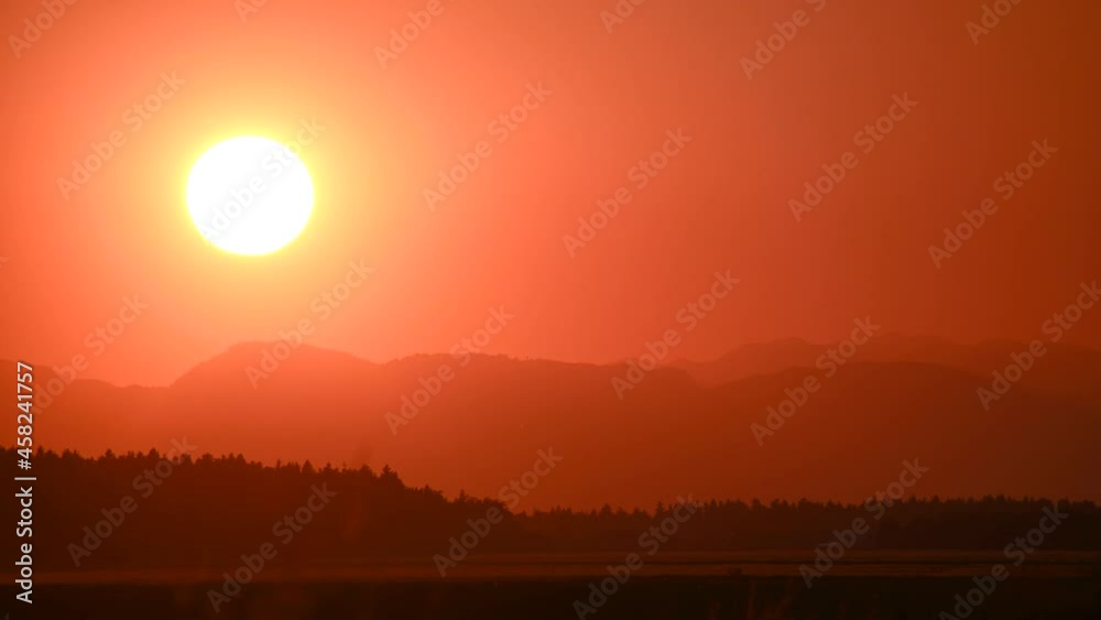 Scenic telephoto perspective of big sun setting behind the mountain. Yellow and orange sunset. Huge bright yellow sun moves down. Silhouette of forest and hills in the distance. Real time, static shot Scenic telephoto perspective of big sun setting behind the mountain. Yellow and orange sunset. Huge bright yellow sun moves down. Silhouette of forest and hills in the distance. Real time, static shot