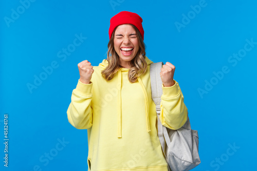 Happy cheerful blond girl in red beanie and yellow hoodie, close eyes relieved and smiling triumphing, celebrating great news, passed exams in university, fist pump like champion, winning prize photo