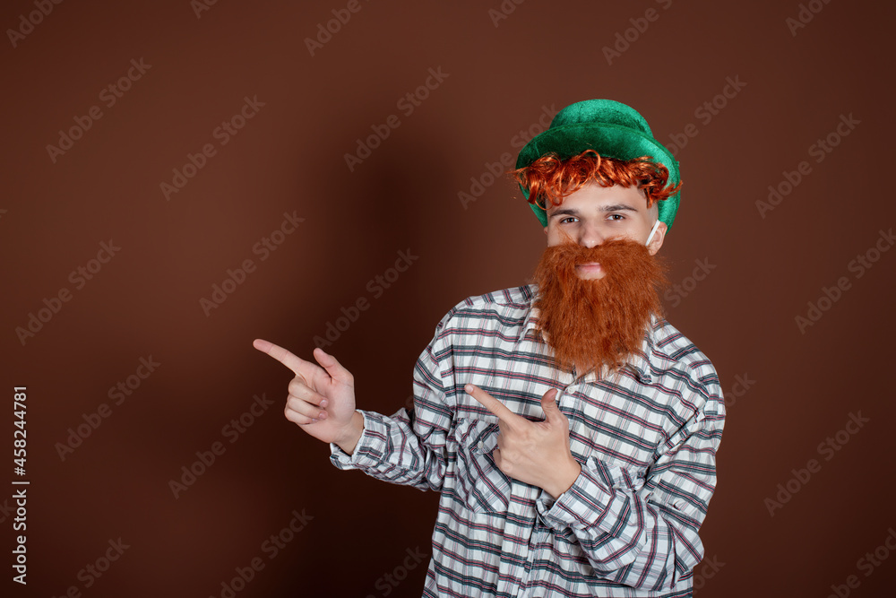 A funny guy in a plaid shirt and a leprechaun hat shows hand gestures. The man on a brown background. Celebration and tradition.