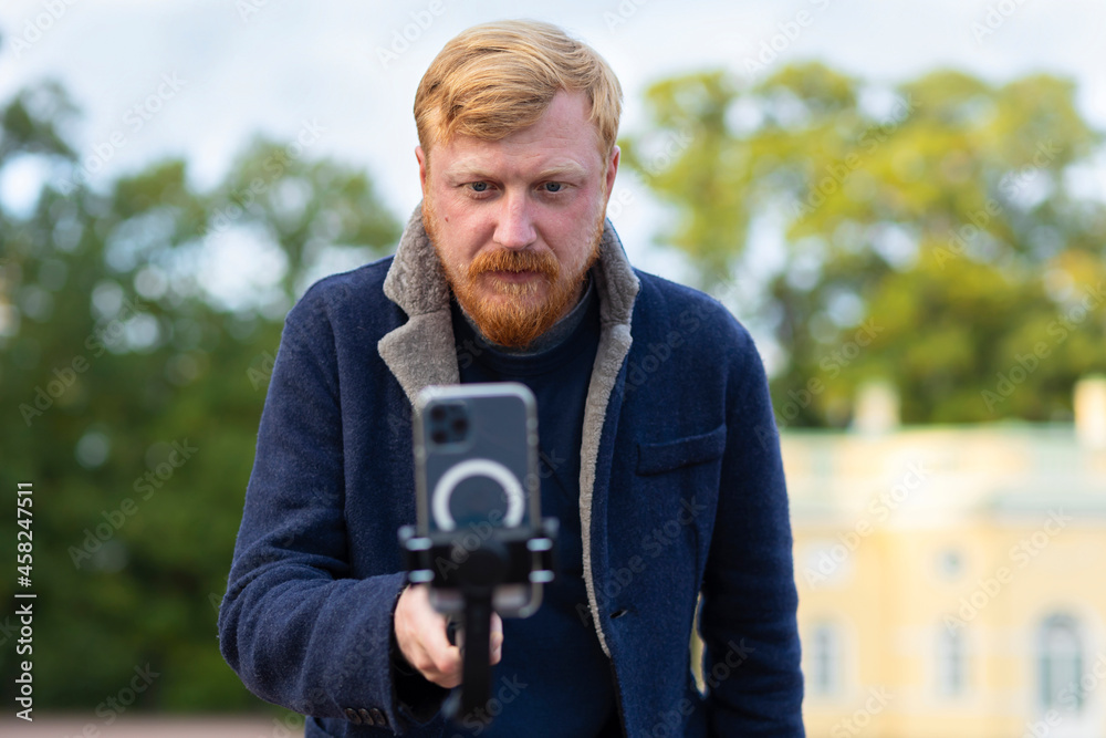 red haired photographer, videographer man with beard, mustache taking story, photo, video smartphone, mobile. stylish ginger bearded guy using phone on selfie stick with stabilizer outdoor red haired photographer, videographer man with beard, mustache taking story, photo, video smartphone, mobile. stylish ginger bearded guy using phone on selfie stick with stabilizer outdoor