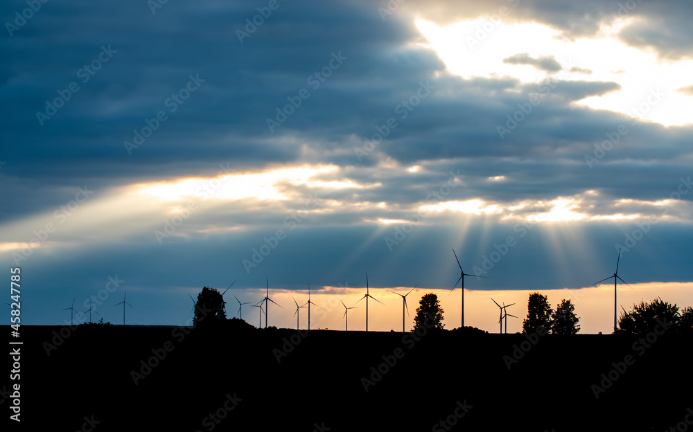 Silhouette of wind turbines and trees on field at the horizon. Dramatic glowing sky with clouds during sunset. 