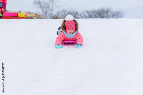 Funny little toddler girl in pink jumpsuit sledding down the hill. wintertime for children. winter games outdoors. Kid having fun at Christmas time