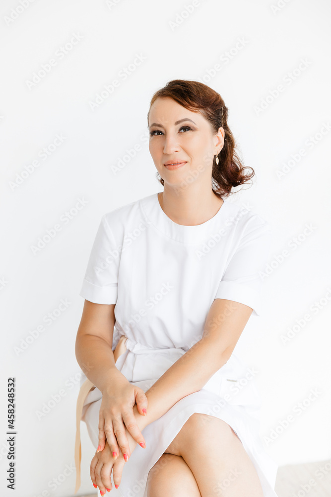 Portrait of a beautiful Caucasian medical girl in a white coat sitting on a stool on a white background. beauty salon. Portrait of a beautiful Caucasian medical girl in a white coat sitting on a stool on a white background. beauty salon.