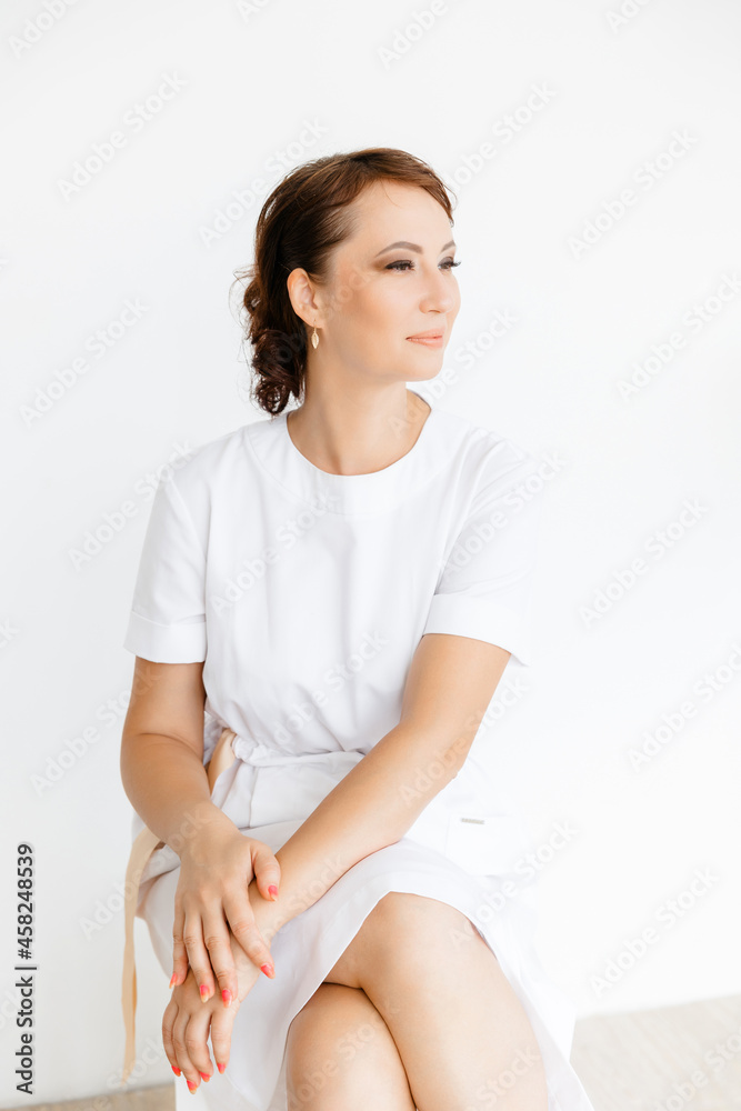 Portrait of a beautiful Caucasian medical girl in a white coat sitting on a stool on a white background. beauty salon. Portrait of a beautiful Caucasian medical girl in a white coat sitting on a stool on a white background. beauty salon.