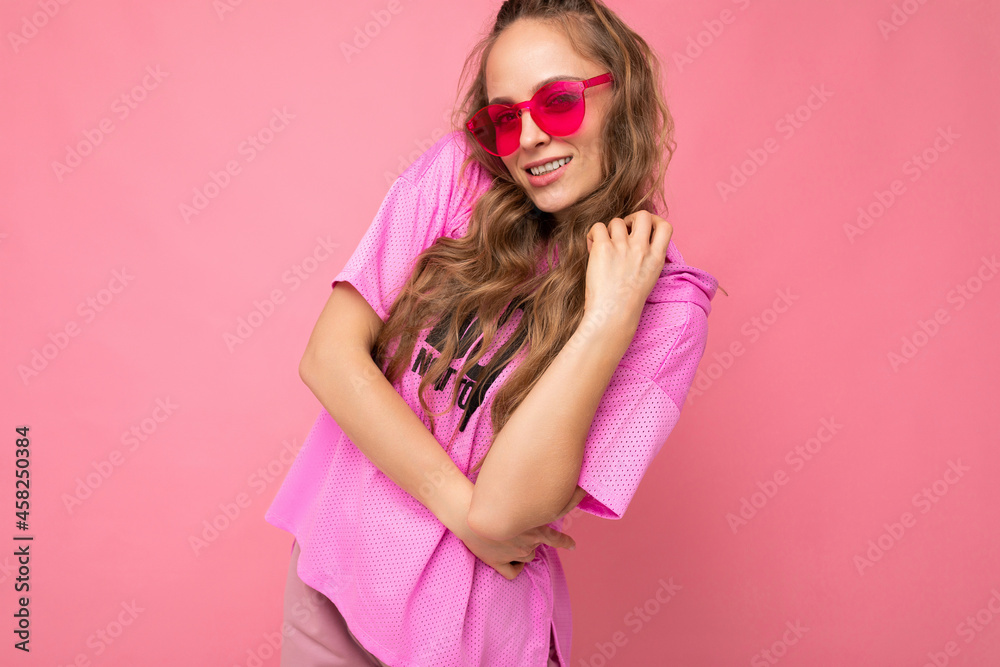 Pretty positive sexy young blonde curly woman isolated over pink background wall wearing casual pink t-shirt and stylish colourful sunglasses looking at camera