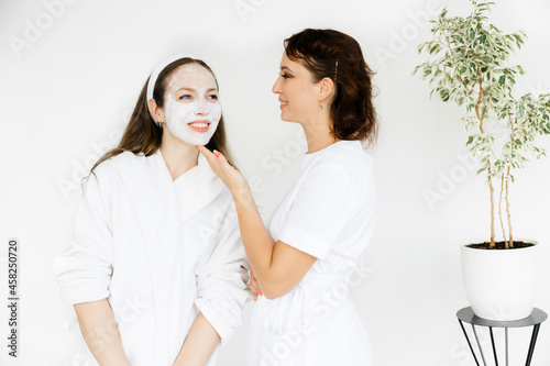 caucasian girl in a white coat with a white mask on her face. the cosmetologist is standing nearby. Facial peeling mask, spa beauty treatments, skin care.