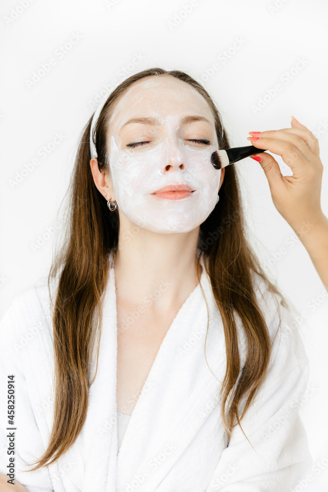 caucasian girl in a white coat with a white mask on her face. the cosmetologist applies the mask to the face with a brush. Facial peeling mask, spa beauty treatments, skin care. caucasian girl in a white coat with a white mask on her face. the cosmetologist applies the mask to the face with a brush. Facial peeling mask, spa beauty treatments, skin care.