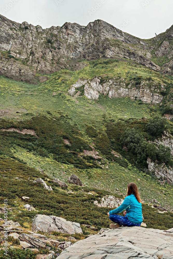 Fototapeta premium rear view of meditating young woman in blue sportswear sitting on stone in green mountain valley, relaxation in nature, consciousness, mindfulness
