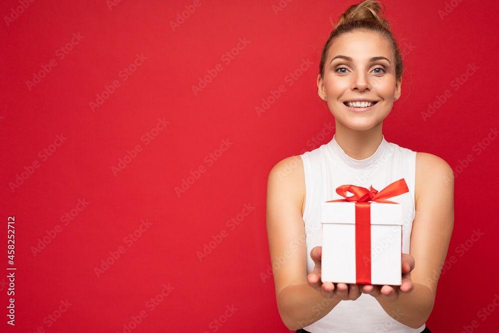 Shot of attractive positive smiling young blonde woman isolated over colourful background wall wearing everyday trendy outfit holding gift box and looking at camera Shot of attractive positive smiling young blonde woman isolated over colourful background wall wearing everyday trendy outfit holding gift box and looking at camera