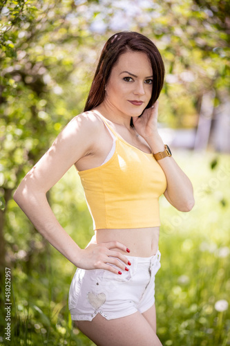 Young woman in yellow top and white shorts standing under trees in summer