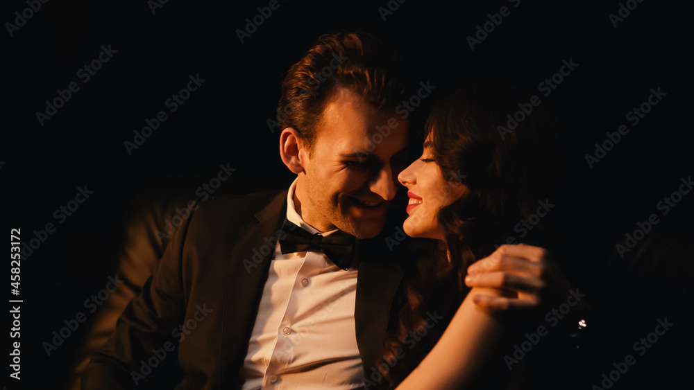 Obraz premium Smiling man in suit hugging and flirting with cheerful girlfriend isolated on black