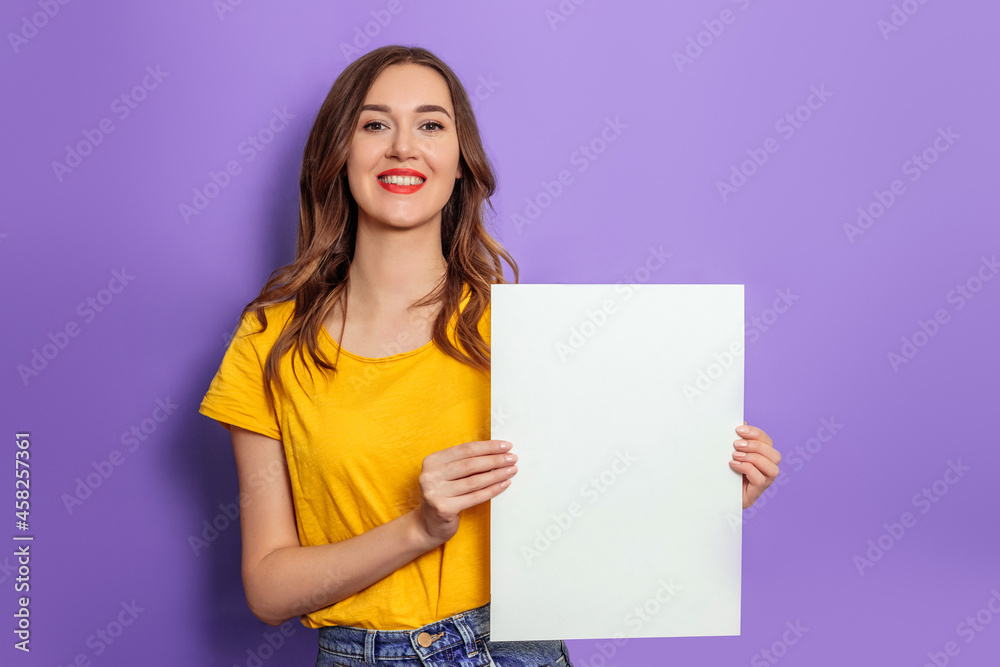Caucasian young woman smiling and holding blank poster wearing yellow t-shirt isolated over lilac background in studio. Mockup for design Caucasian young woman smiling and holding blank poster wearing yellow t-shirt isolated over lilac background in studio. Mockup for design