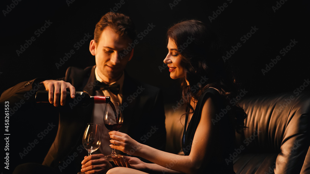 Happy man pouring wine near cheerful woman holding glass isolated on black