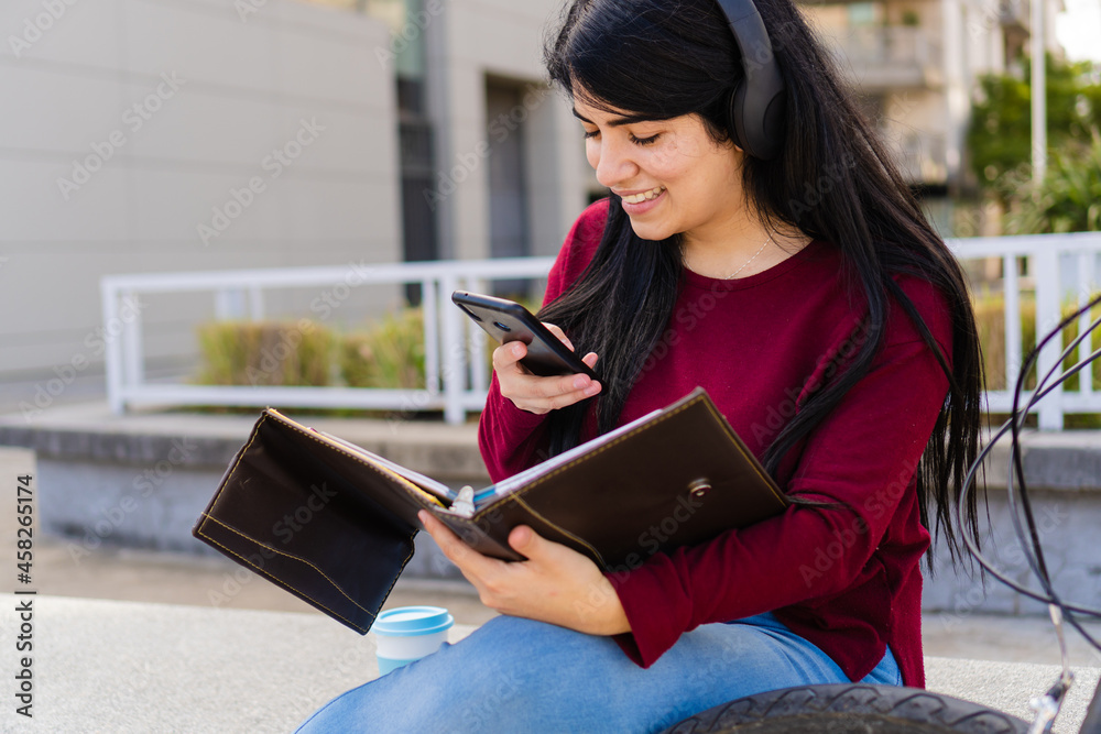 Fototapeta premium Young latin woman taking a picture to her notes of notebook outdoors