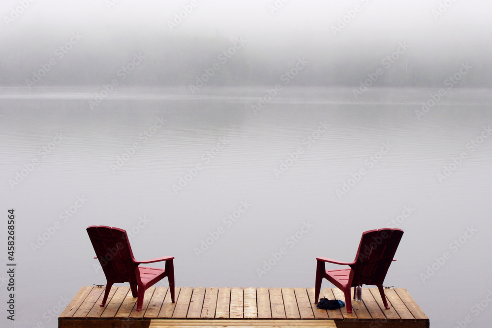 two Adirondack chairs on wooden deck at edge of lake in fog with copy space