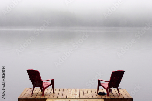 two Adirondack chairs on wooden deck at edge of lake in fog with copy space