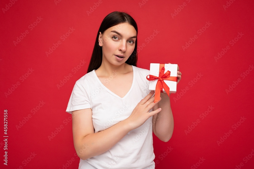 Photo of attractive positive amazed young brunet woman isolated over red background wall wearing white t-shirt for mockup holding white gift box with red ribbon and looking at camera