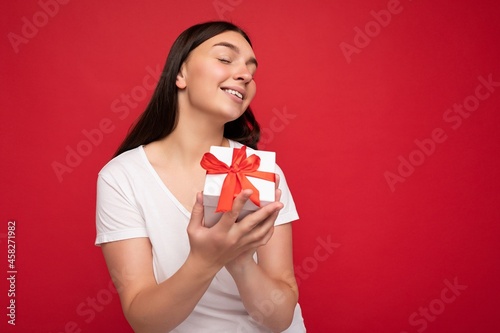 Beautiful happy young brunette woman isolated over colourful background wall wearing stylish casual clothes holding gift box and looking to the side