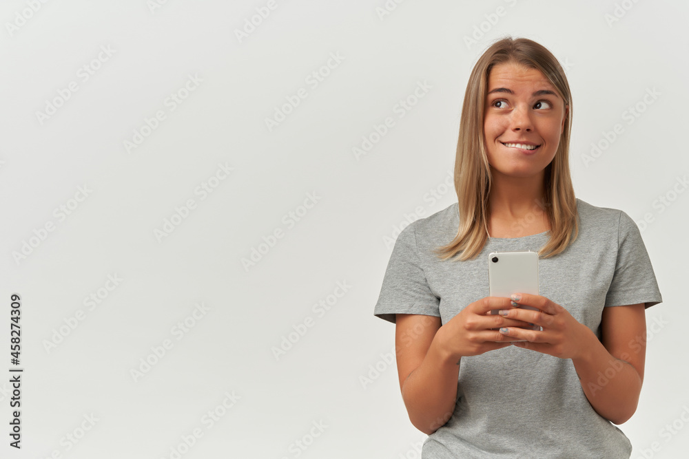 Indoor shot of young adult female, wears gray t-shirt, texting with her boyfriend, looks aside and biting her lip, smiles broadly. Isolated over white background Indoor shot of young adult female, wears gray t-shirt, texting with her boyfriend, looks aside and biting her lip, smiles broadly. Isolated over white background