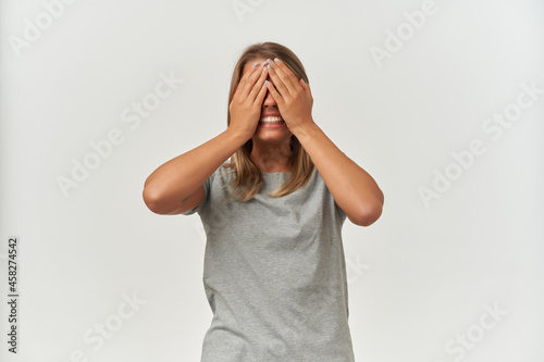 Young playful female, wears gray t-shirt, closed her eyes with palms and smiles broadly. Isolated over white background