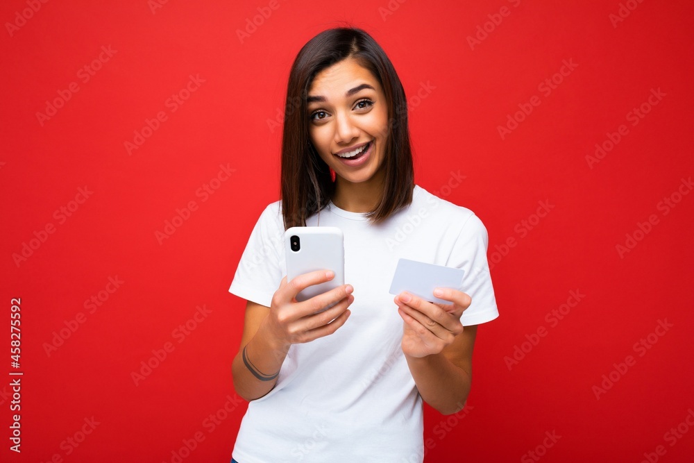 Young smiling happy young woman wearing everyday clothes isolated over background holding phone and credit card paying online shopping through credit card looking at camera Young smiling happy young woman wearing everyday clothes isolated over background holding phone and credit card paying online shopping through credit card looking at camera