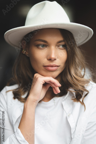 Stylish girl in white trousers and white cotton blouse posing against the wall. Photo of a woman in a hat.