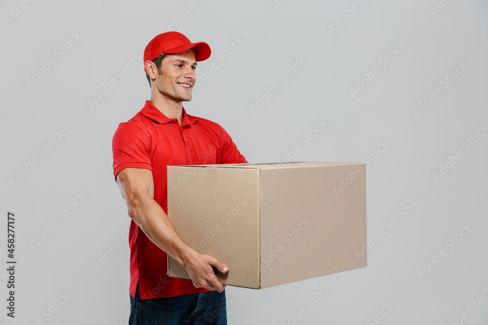 Young delivery man in hat smiling while posing with cardboard box Young delivery man in hat smiling while posing with cardboard box