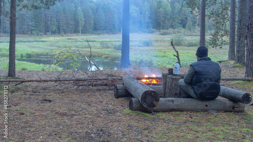 a camping camp on the river bank, a bonfire is burning nearby. A man sits on a log next to a fire.