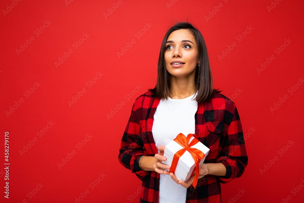 Photo shot of pretty positive young brunette woman isolated over red background wall wearing white casual t-shirt and red and black shirt holding white gift box with red ribbon and looking up Photo shot of pretty positive young brunette woman isolated over red background wall wearing white casual t-shirt and red and black shirt holding white gift box with red ribbon and looking up
