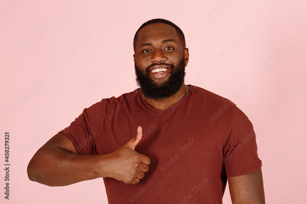 Smiling bearded Afro-American guy in stylish brown t-shirt shows thumb-up standing on pink background in studio close view Smiling bearded Afro-American guy in stylish brown t-shirt shows thumb-up standing on pink background in studio close view