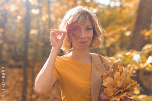 Young blonde woman covering one eye with red maple leaf. Autumn and season concept. Outdoor fall female portrait close-up with foliage