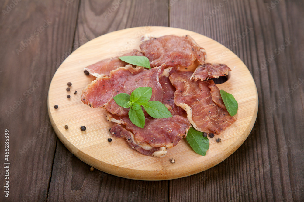 Pieces of jerky meat with basil leaves lie on a wooden cutting board on a wooden background.