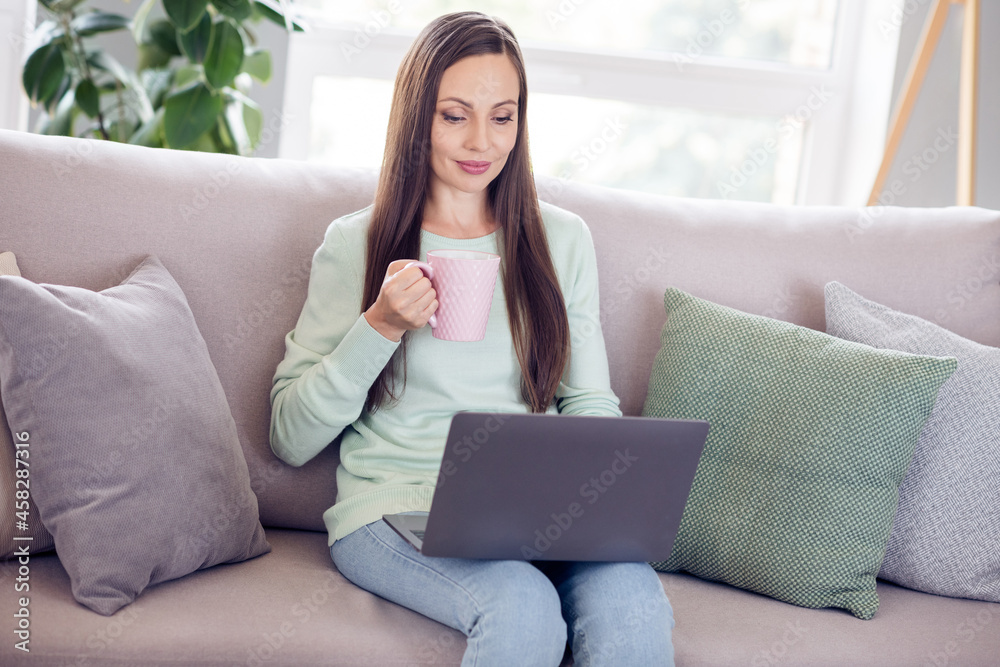 Photo of charming cute young woman dressed turquoise sweater sitting sofa drinking tea looking modern device indoors home room Photo of charming cute young woman dressed turquoise sweater sitting sofa drinking tea looking modern device indoors home room