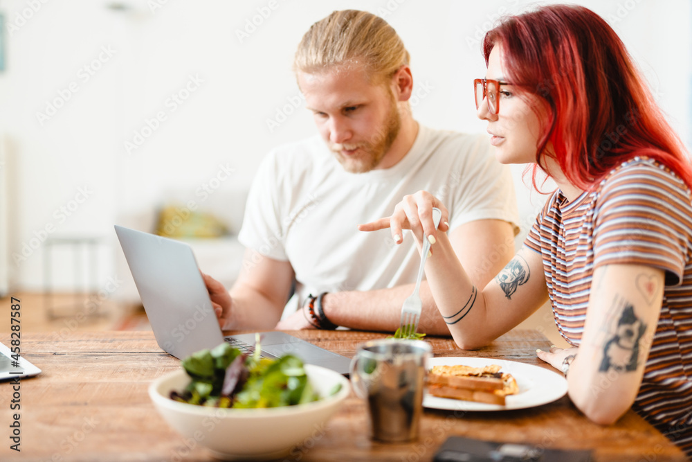 Young white couple using laptop and having lunch while sitting at table Young white couple using laptop and having lunch while sitting at table