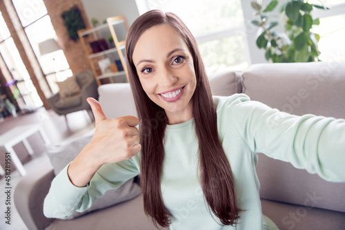 Photo of shiny sweet mature lady wear teal pullover smiling sitting couch recording video showing thumb up indoors house room