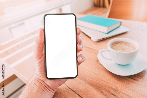 Blank mobile smart phone screen mock up. Man holding smartphone over desk with coffee cup at home.