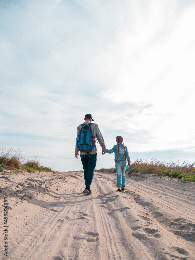 Happy father and daughter running jumping having fun on empty autumn sea beach. Dad and child walking on white sand road with reeds blue sky background. Lifestyle real people. Happiness travel concept