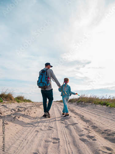 Happy father and daughter running jumping having fun on empty autumn sea beach. Dad and child walking on white sand road with reeds blue sky background. Lifestyle real people. Happiness travel concept