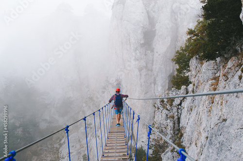 A young man in mountaineering equipment walks uphill on a suspension bridge in the fog. Suspension bridge on Mount Ai-Petri, Yalta, Crimea. The concept of travel, active lifestyle, tourism. photo