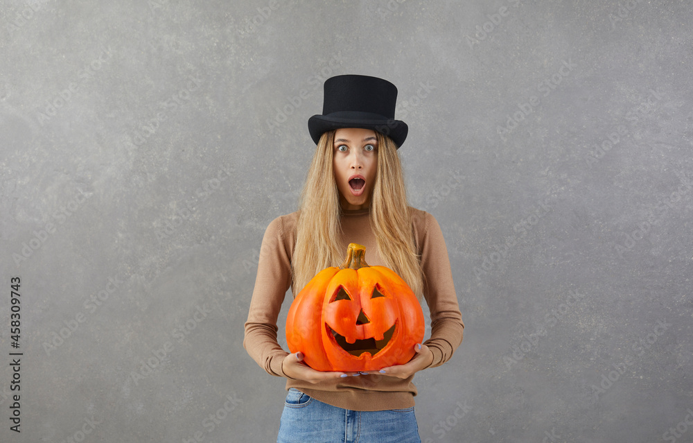 © Studio Romantic - Woman in black top hat with modern festive orange Jack-o-lantern pumpkin standing against grey studio background looking at camera with surprised, shocked or scared face expression. Halloween concept