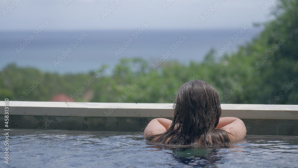 A white girl with long dark loose hair is resting on the edge of the pool with a wonderful view of the blue ocean and the sky. A woman gets a tan in the pool and looks into the sea on a sunny day. A white girl with long dark loose hair is resting on the edge of the pool with a wonderful view of the blue ocean and the sky. A woman gets a tan in the pool and looks into the sea on a sunny day.