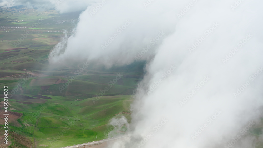 A sky shot shot through rain clouds with a view of green and brown earth fields. A bird's-eye view above the clouds with a view of the road and a rural valley on a sunny day. Aerial copter frame. A sky shot shot through rain clouds with a view of green and brown earth fields. A bird's-eye view above the clouds with a view of the road and a rural valley on a sunny day. Aerial copter frame.