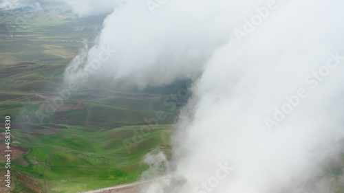 A sky shot shot through rain clouds with a view of green and brown earth fields. A bird's-eye view above the clouds with a view of the road and a rural valley on a sunny day. Aerial copter frame.