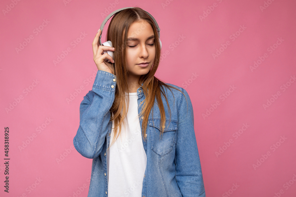 Attractive with sincere emotions young blonde woman wearing blue jean shirt and white t-shirt isolated over pink background wearing white wireless bluetooth headphones listening to cool music and Attractive with sincere emotions young blonde woman wearing blue jean shirt and white t-shirt isolated over pink background wearing white wireless bluetooth headphones listening to cool music and